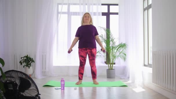 Fitnesstraining - blonde übergewichtige Frau macht Fitnessübungen - steht auf Yogamatte und trainiert ihre Hände mit kleinen Hanteln — Stockvideo