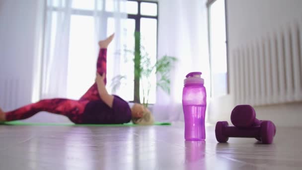 健身训练- -超重的女人仰卧在床上，为腿做伸展运动- -前景是瓶子和小哑铃 — 图库视频影像