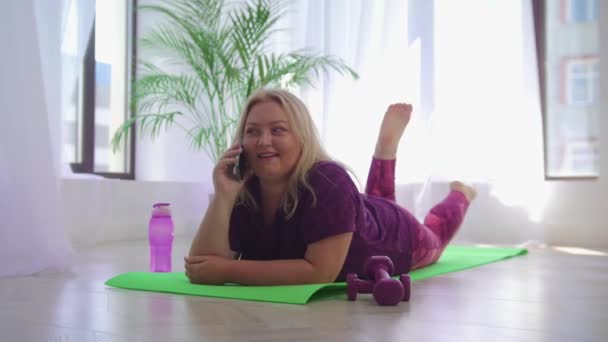 Улыбающаяся толстуха лежит на коврике для йоги и разговаривает по телефону. — стоковое видео