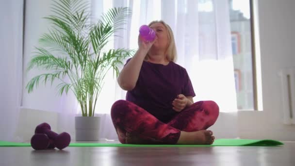 Фитнес-тренировка - блондинка с избыточным весом, сидящая на коврике для йоги и пьющая воду из бутылки — стоковое видео