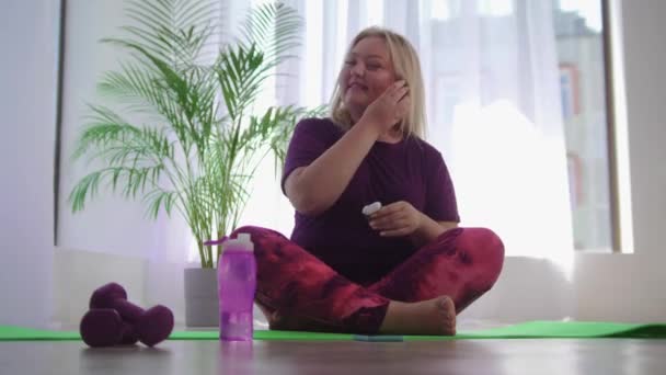 Тренировка фитнеса - блондинка с избыточным весом сидит на коврике для йоги и надевает беспроводные наушники — стоковое видео