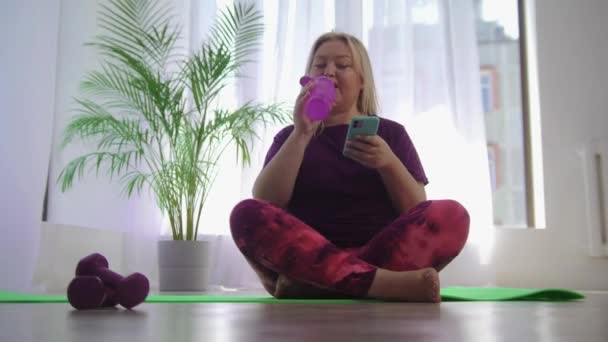 Fitness školení - blondýny nadváha žena sedí na podložce jógy a dívá se na telefon při pití vody z láhve — Stock video