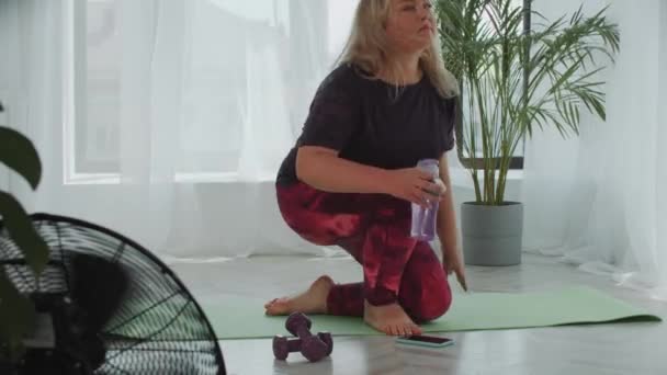 Blond overgewicht vrouw gaat zitten op yoga mat en zet in draadloze hoofdtelefoon zet de muziek op voor de training — Stockvideo