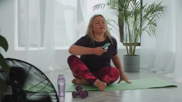 Blonde femme en surpoids s'assoit sur un tapis de yoga et met des écouteurs sans fil avant la formation — Video