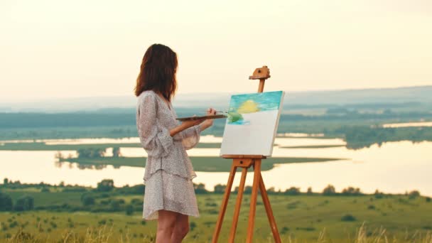 Mujer joven con cabello oscuro dibujando una pintura al atardecer y mira a la cámara — Vídeo de stock