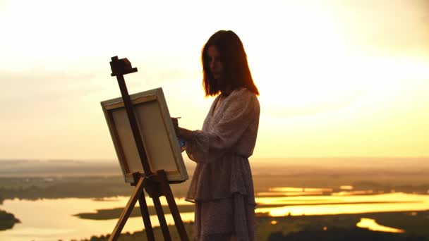 Egy fiatal, gyönyörű nő rajzol egy festményt a naplemente mezőn. — Stock videók