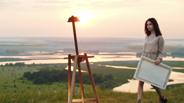 Seorang seniman wanita muda berbusana terang berjalan ke sebuah sela-sela yang berdiri di lapangan matahari terbenam dan meletakkan sebuah kanvas di atasnya — Stok Video