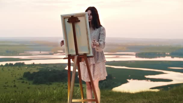 Een jonge kunstenares wandelt een doek op een ezel op een zonsondergang veld en kijkt uit over het landschap — Stockvideo