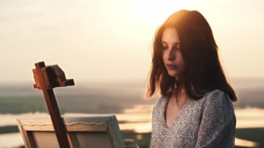 Yüzünde çiller olan genç ve güzel bir kadın sanatçı gün batımında bir resim çizer.