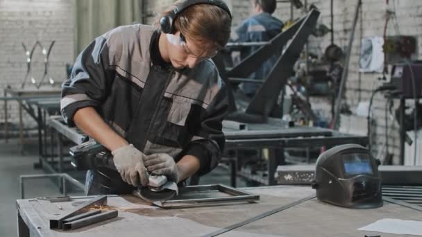 Młody człowiek pracujący w zakładzie - szlifowanie konstrukcji metalowej szlifierką kątową — Wideo stockowe