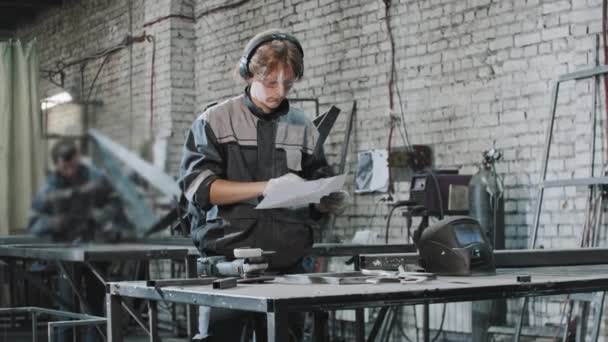 Νεαρός άνδρας με προστατευτικά γυαλιά που εργάζεται στο εργοστάσιο - ιππεύοντας τις οδηγίες στο χαρτί — Αρχείο Βίντεο