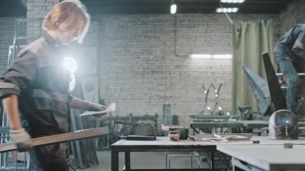 El joven que trabaja en la planta trae detalles largos de metal y la instrucción a la mesa y la lectura de la instrucción — Vídeo de stock