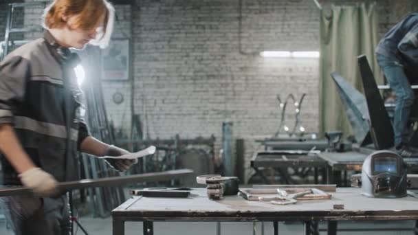 Jongeman met lang haar aan het werk in de fabriek - brengt metaal lang detail en instructies aan de tafel en het lezen van de instructie — Stockvideo