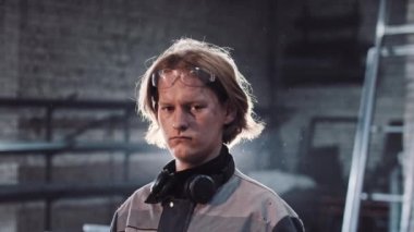 Fabrikada çalışan genç bir adam. Elinde açı öğütücüsü var ve kameraya bakıyor.