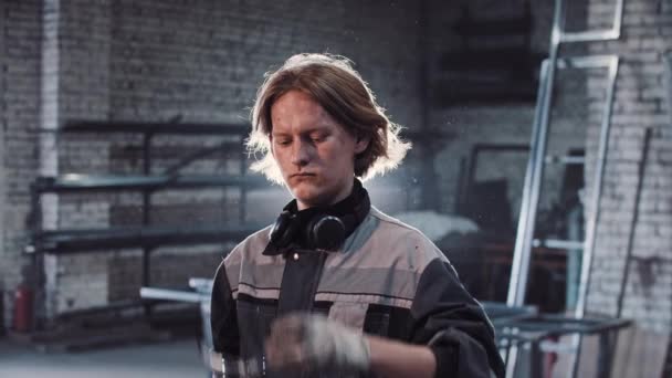 Молодий чоловік з довгим волоссям, що працює на заводі - тримає кутову шліфувальну машину і дивиться в камеру — стокове відео