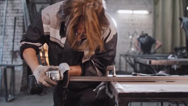 Młody człowiek z długimi włosami pracujący w zakładzie budowlanym - szlifowanie detali metalowych — Wideo stockowe