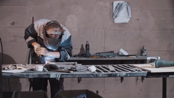 Młody pracownik w rękawiczkach ochronnych i hełmach spawalniczych części metalowych detali razem przy stole — Wideo stockowe