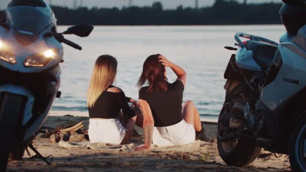 Due donne amiche motociclisti seduti sulla spiaggia e guardando l'acqua a tarda sera del tramonto — Video Stock