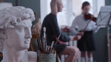 Genç bayan öğretmen küçük bir kıza keman dersi veriyor - Yunan adam ön planda büst ve sanat fırçaları