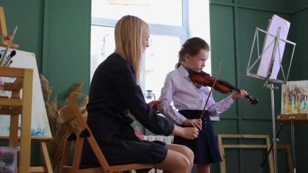 バイオリン教室-小さな女の子再生バイオリンでザクラスと彼女のブロンド女性先生座っています彼女の隣にと固定彼女の位置 — ストック動画