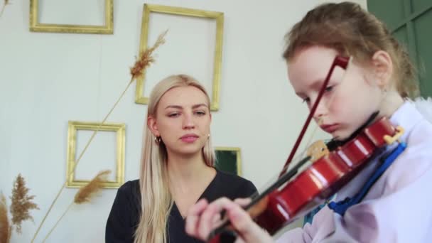 Μάθημα βιολιού - ένα μικρό κορίτσι που παίζει βιολί με ένα τόξο στην τάξη και ο δάσκαλός της κάθεται δίπλα της και μιλάει — Αρχείο Βίντεο