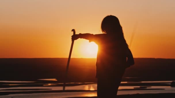 Mujer joven en una sudadera con capucha balanceándose con dos espadas al atardecer naranja tardío — Vídeo de stock