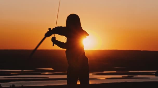Fiatal nő kapucnis pulcsiban, két karddal a szabadban, késő narancssárga naplementében. — Stock videók