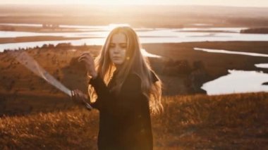 Siyah elbiseli genç bir kadın doğada günbatımında kılıçla antrenman yapıyor.