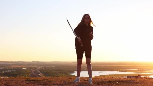Svärd utbildning - ung kvinna med långt hår viftar med ett svärd i cirklar på bakgrunden av den ljusa solnedgången — Stockvideo