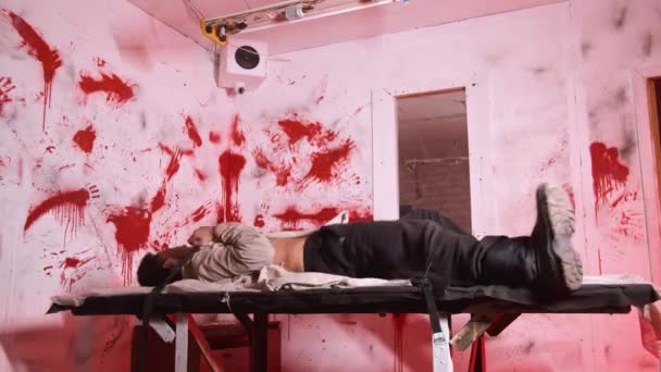 Horror acting - an aggressive man tied up to the table in bloody room of mental hospital and trying to get out — Stock Video