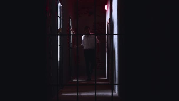 Horror actuando - un peligroso guardia masculino camina por las celdas de la prisión sosteniendo una tubería de metal y golpea las manos del prisionero en las barras con una tubería — Vídeos de Stock