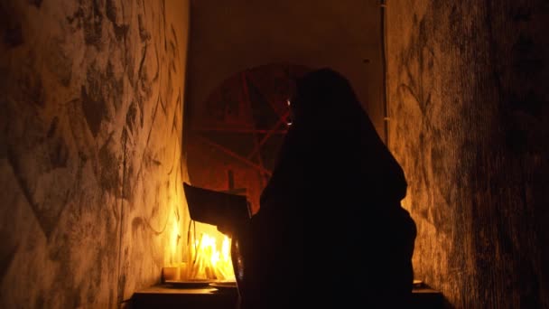 A man occultist in black robes stands with a candle in his hand and reading a book — Stock Video