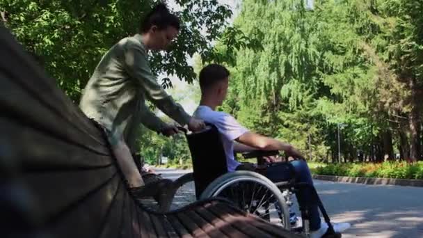 Een vrouw brengt een man in een rolstoel naast een bank en zit in de buurt — Stockvideo