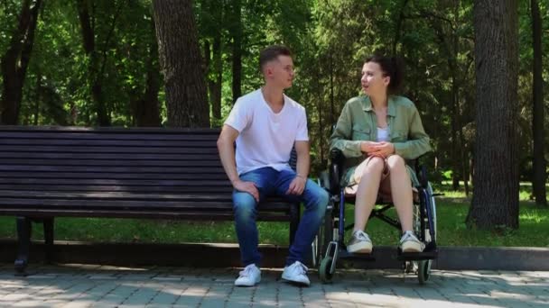 Le type accompagne une femme en fauteuil roulant et lui parle tout en étant assis à côté d'elle sur un banc — Video