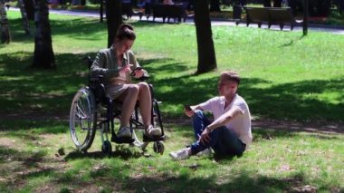 Adam tekerlekli sandalyede bir kadına eşlik ediyor. Parkta oturuyor ve telefonlarına bakıyor. Adam bir kadına telefonundan bir resim gösteriyor.