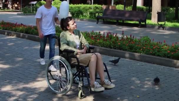 Bored woman in a wheelchair sitting in the park - her boyfriend comes to her from the back and brings to her a cotton candy — Stock Video