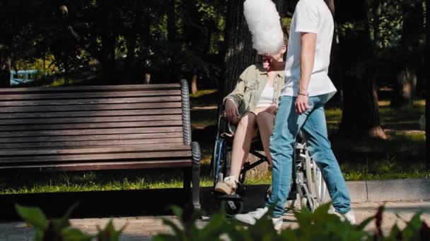 一个年轻人把棉花糖拿给公园里坐轮椅的女人，然后坐在长椅上 — 图库视频影像