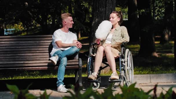Een jonge man zit op een bank en een vrouw in een rolstoel eet suikerspin naast zich. — Stockvideo