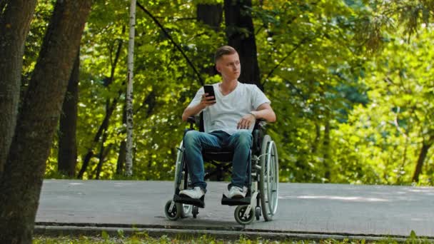 Een man in een rolstoel zittend aan zijn telefoon op een voetgangerspad — Stockvideo