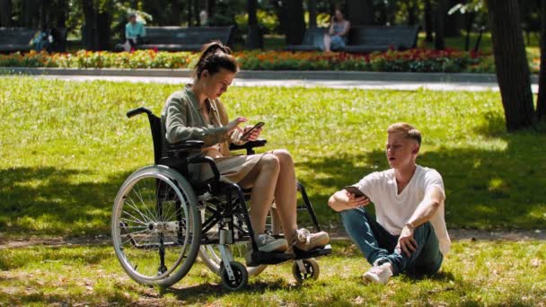 Mulher em uma cadeira de rodas e seu amigo passar o tempo no parque de verão e sentado em seus telefones — Vídeo de Stock