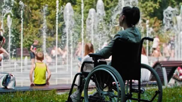 Mujer triste en una silla de ruedas sentada frente a una fuente y mirando a la gente feliz — Vídeos de Stock