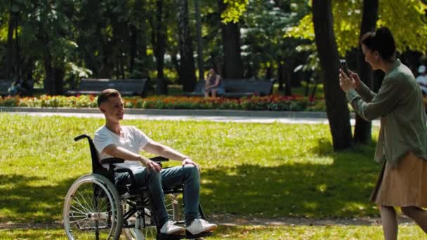 Hombre en silla de ruedas y su amiga pasando tiempo en el parque de verano y la mujer tomando una foto de él el teléfono — Vídeos de Stock