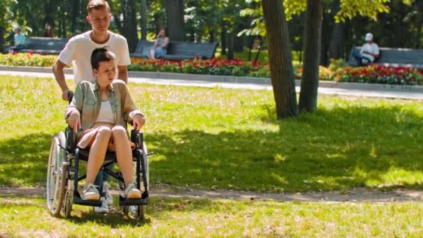 Vrienden in het park - een man rijdt gehandicapte vrouw in een rolstoel dan stopt en zit naast haar op de grond — Stockvideo