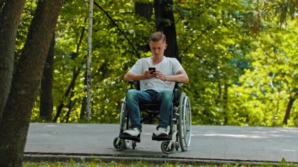 Een man in een rolstoel zit achter zijn telefoon op een voetgangerspad en staat op het punt weg te rijden. — Stockvideo