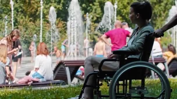 El hombre trae a la mujer en una silla de ruedas para mirar la fuente y la gente en el parque — Vídeo de stock