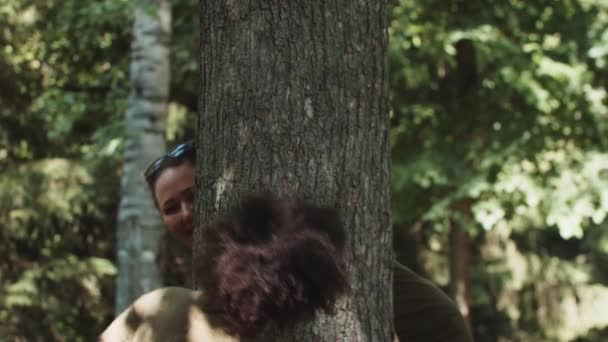 Piccola ragazza nera e la sua madre bianca sorridente che gioca nel parco verde guardare fuori da dietro un albero — Video Stock