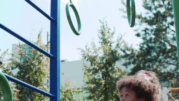 Blanke vrouw en haar zwarte dochtertje spelen op de speelplaats - vrouw helpt een meisje om de hangende ringen te bereiken — Stockvideo