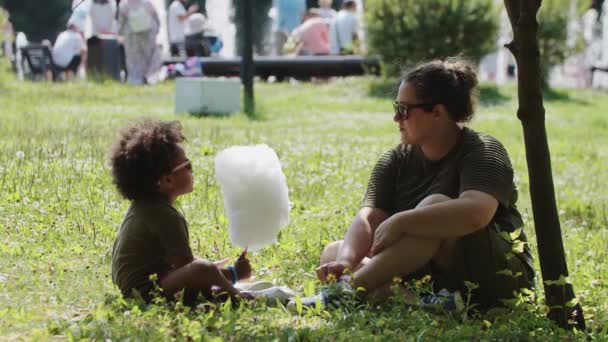 Familia mixta de madre blanca e hija negra que pasan tiempo en el parque comiendo algodón de azúcar sentado en la hierba — Vídeos de Stock