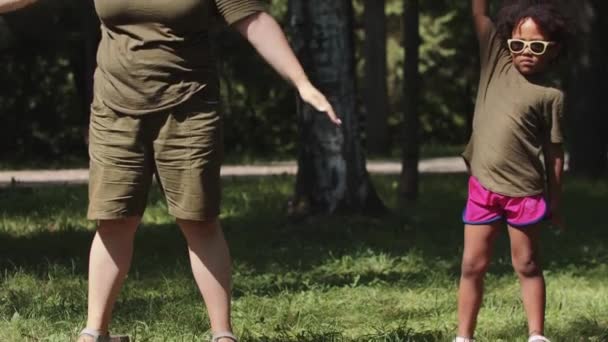 Blanke vrouw en haar zwarte dochtertje in groene kleren doen eenvoudige oefeningen in het park — Stockvideo