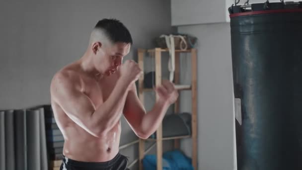 Póló nélküli, izzadt fiatalember bokszoló, aki a boxzsákot veri. — Stock videók